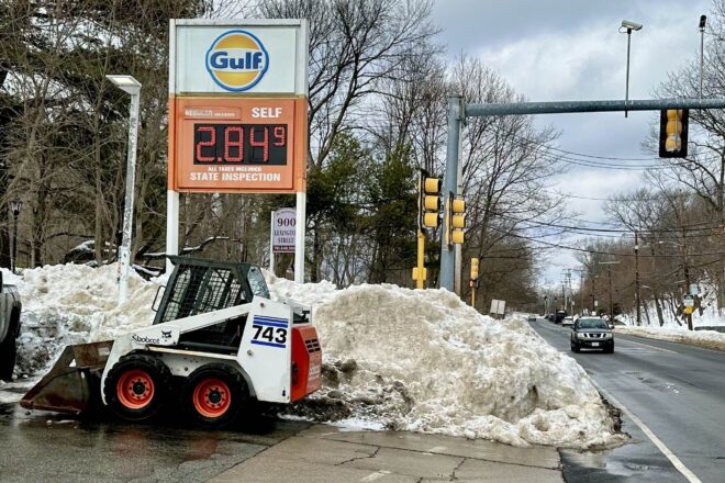 5-foot high snow mound completely blocks sidewalk and shoulder at a gas station, with small plow parked in front of it.