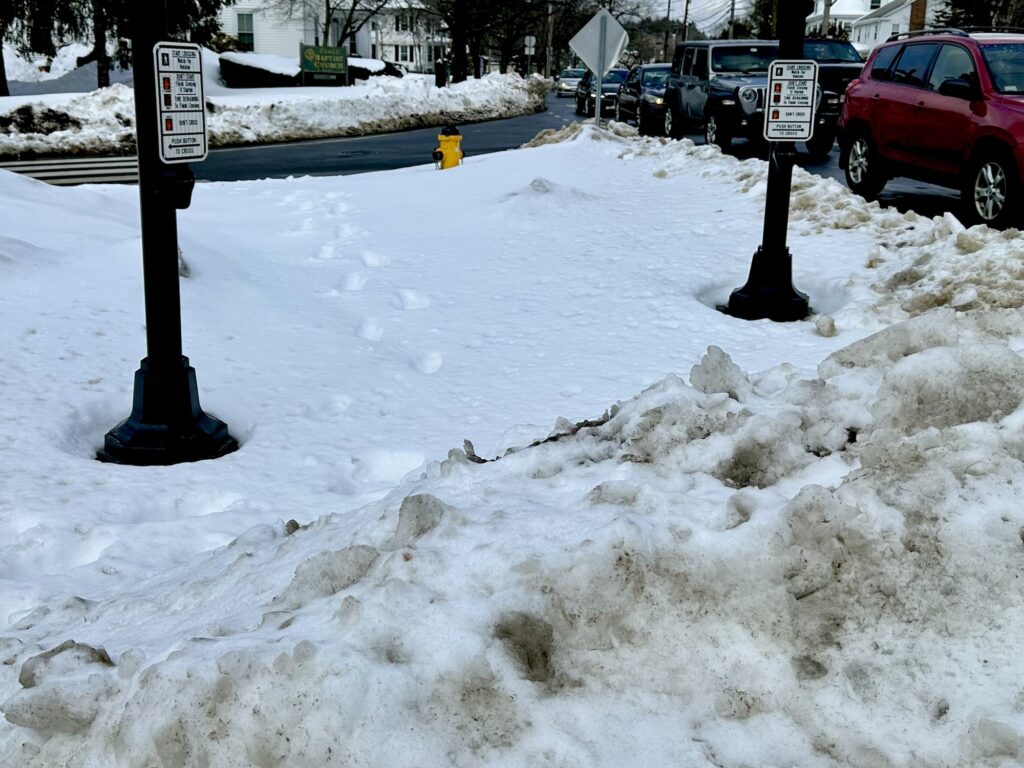 Pedestrian island in the middle of an intersection completely covered in snow, blocking access to the walk signal buttons.